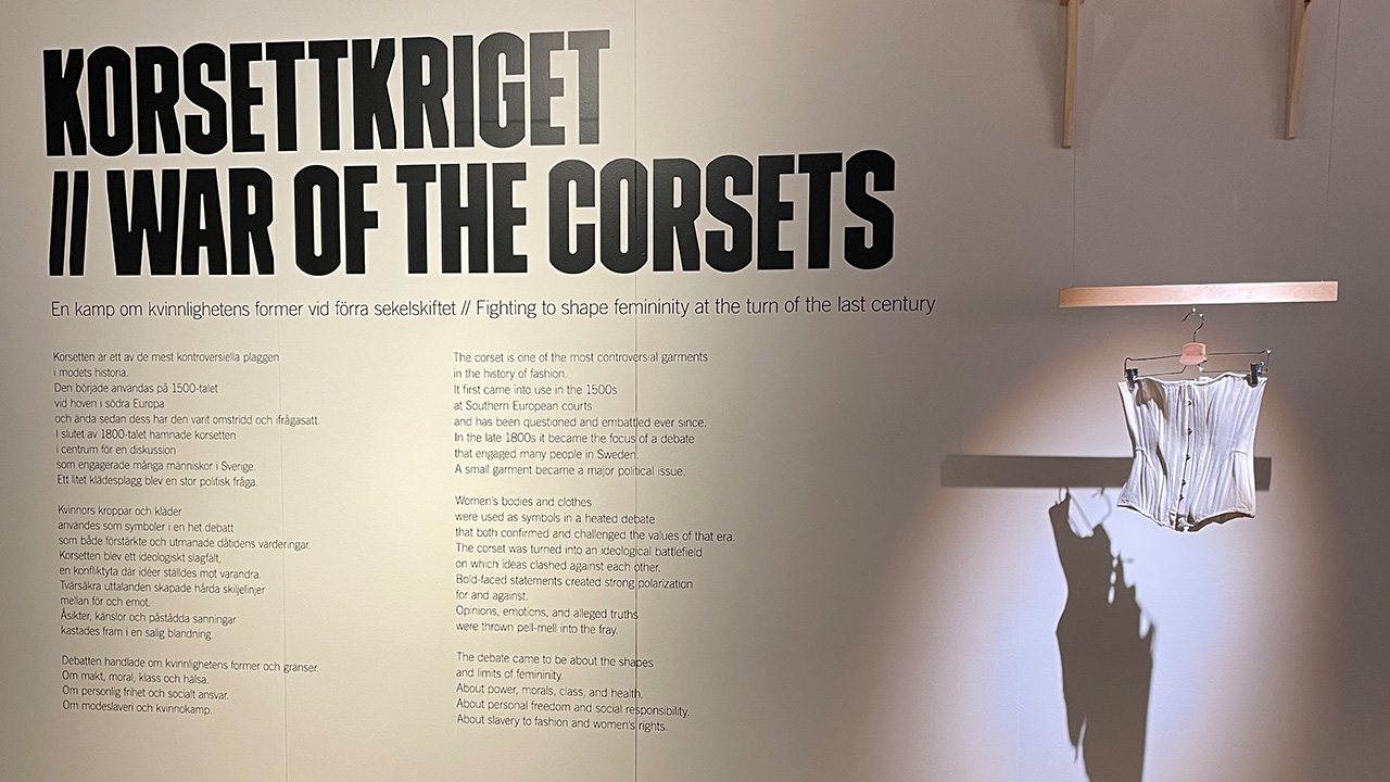 Foto på utställningen Korsettkrigets introtext med en korsett hängande i en träställning till höger. 